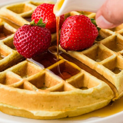 A golden waffle topped with three fresh strawberries is being drizzled with syrup from a white pitcher on a white plate.