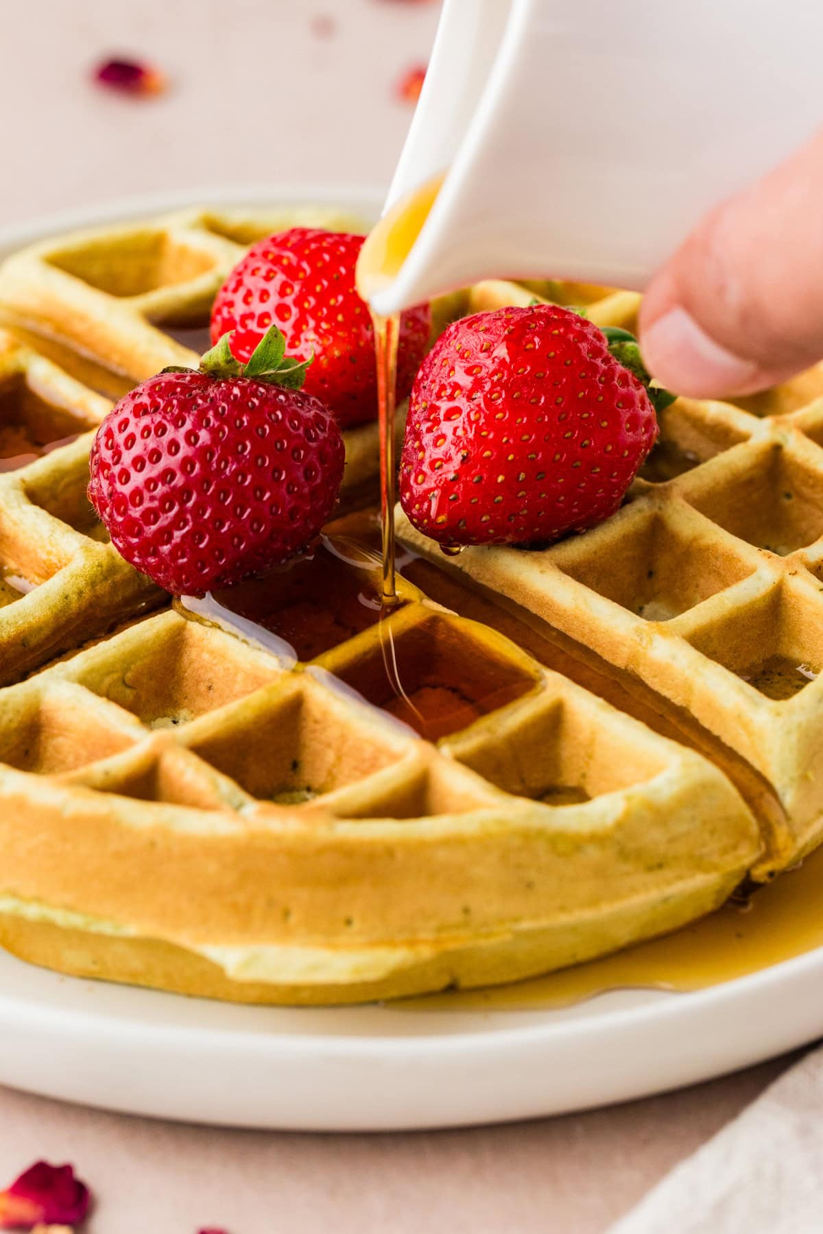 A golden waffle topped with three fresh strawberries is being drizzled with syrup from a white pitcher on a white plate.
