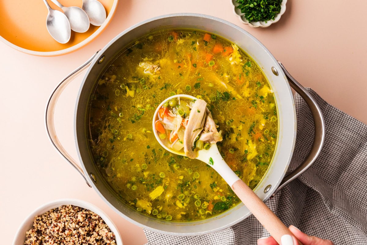 A large pot of chicken soup with vegetables is on a light surface. A hand holds a ladle scooping soup. Nearby are spoons, a bowl of chopped herbs, and a bowl of cooked grains. A checked napkin is partially visible.