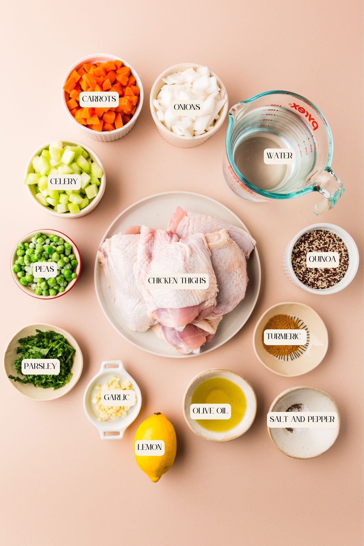 A flat lay of ingredients for a recipe, including raw chicken thighs on a plate, and small bowls containing carrots, onions, celery, peas, parsley, garlic, lemon, olive oil, turmeric, salt and pepper, quinoa, and a measuring cup of water.