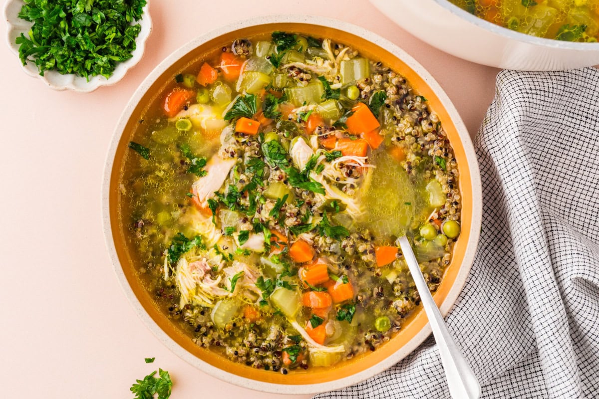 A bowl of chicken and vegetable soup with shredded chicken, carrots, celery, peas, and herbs, garnished with chopped parsley on top, next to a gray and white cloth napkin.