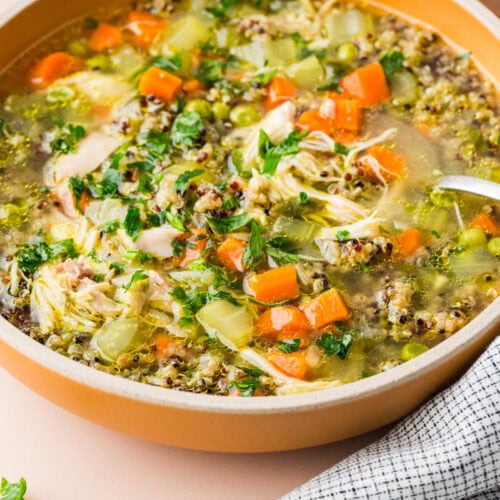 A bowl of hearty chicken and vegetable soup with carrots, celery, shredded chicken, quinoa, and fresh herbs, placed on a light pink surface next to a black and white checkered cloth and a bowl of chopped parsley.