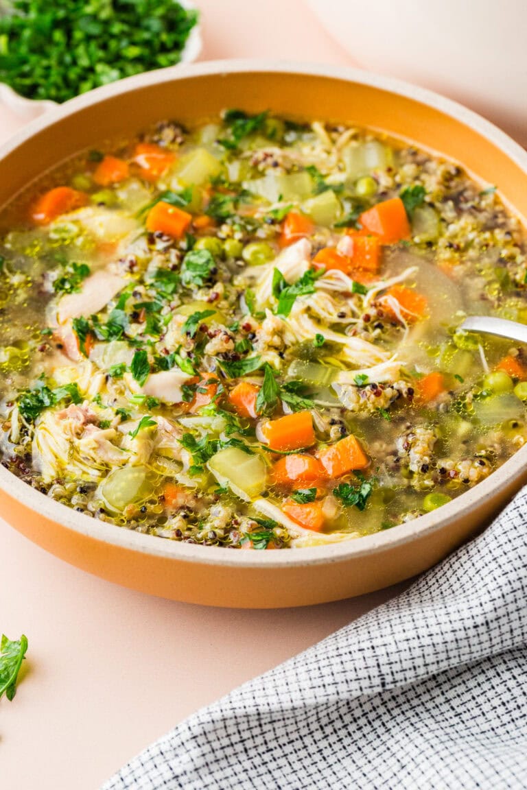 A bowl of hearty chicken and vegetable soup with carrots, celery, shredded chicken, quinoa, and fresh herbs, placed on a light pink surface next to a black and white checkered cloth and a bowl of chopped parsley.