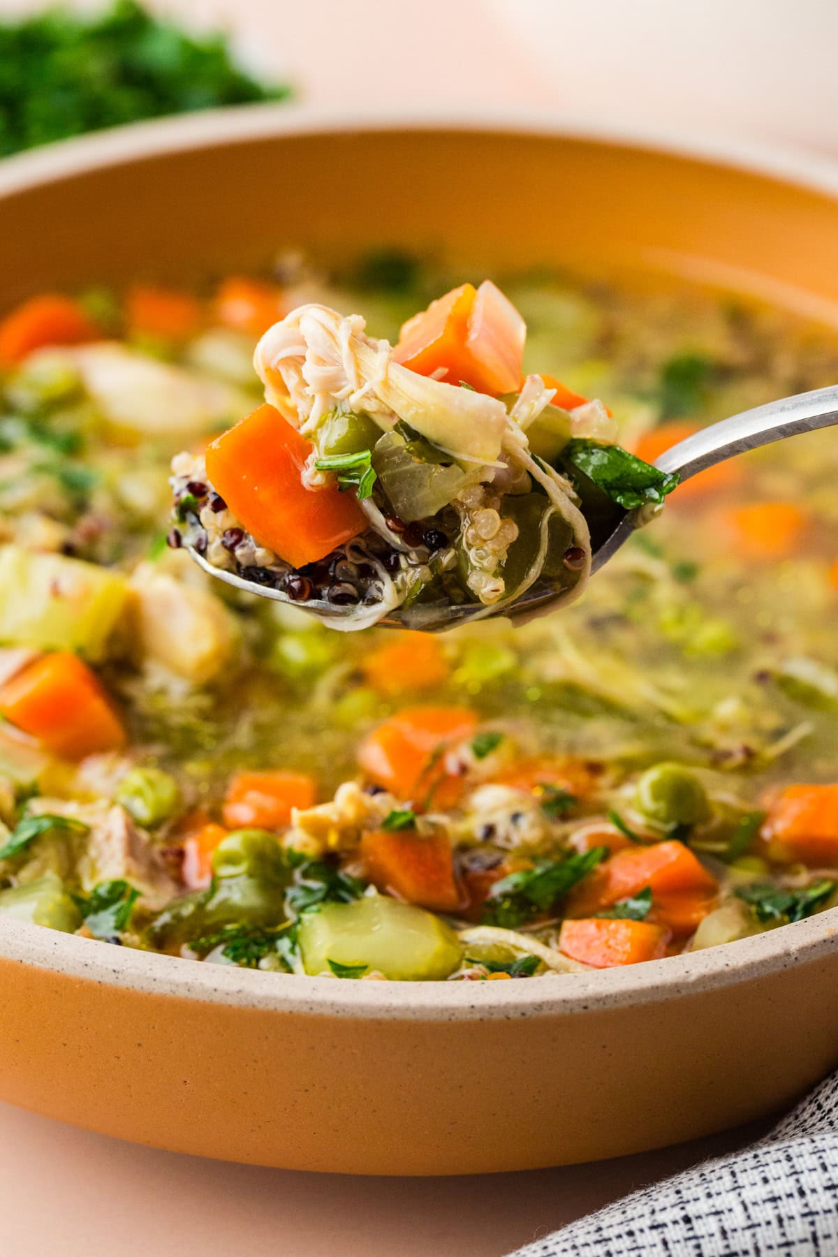 A close-up of a spoonful of vegetable and chicken soup being held over a bowl, showing carrots, celery, greens, and shredded chicken in a clear broth.