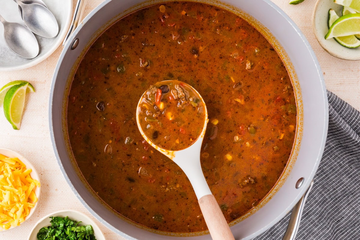 A pot of hearty spicy beef soup with a white ladle lifting a serving. The chunky soup features vegetables, herbs, and tender beef. Slices of lime, shredded cheese, chopped greens, and spoons are arranged nearby.