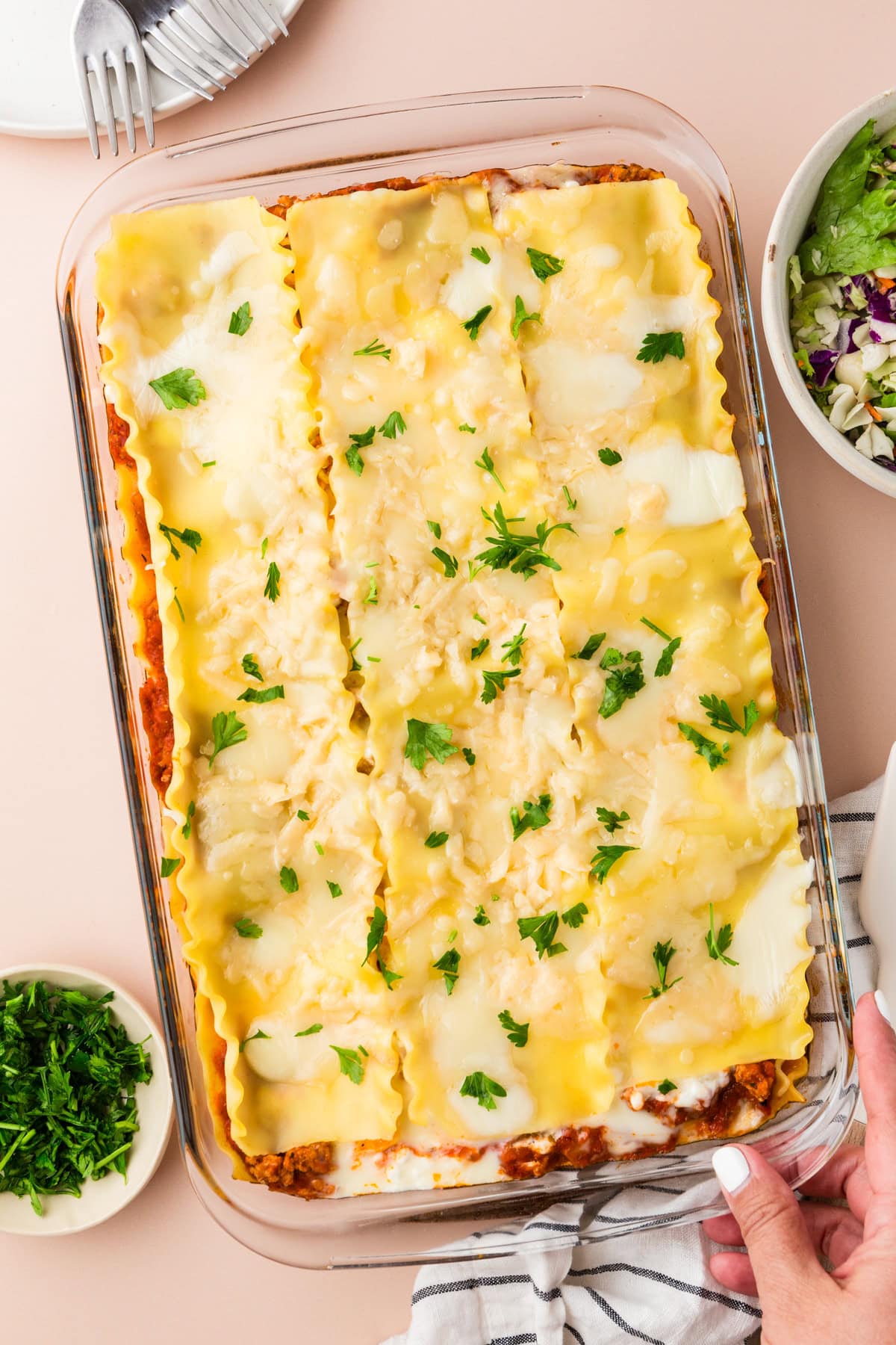 A glass baking dish filled with Ground Turkey Lasagna, topped with melted cheese and parsley. A hand holds the dish on the right. Nearby are bowls of chopped parsley and salad, and a plate with a fork.