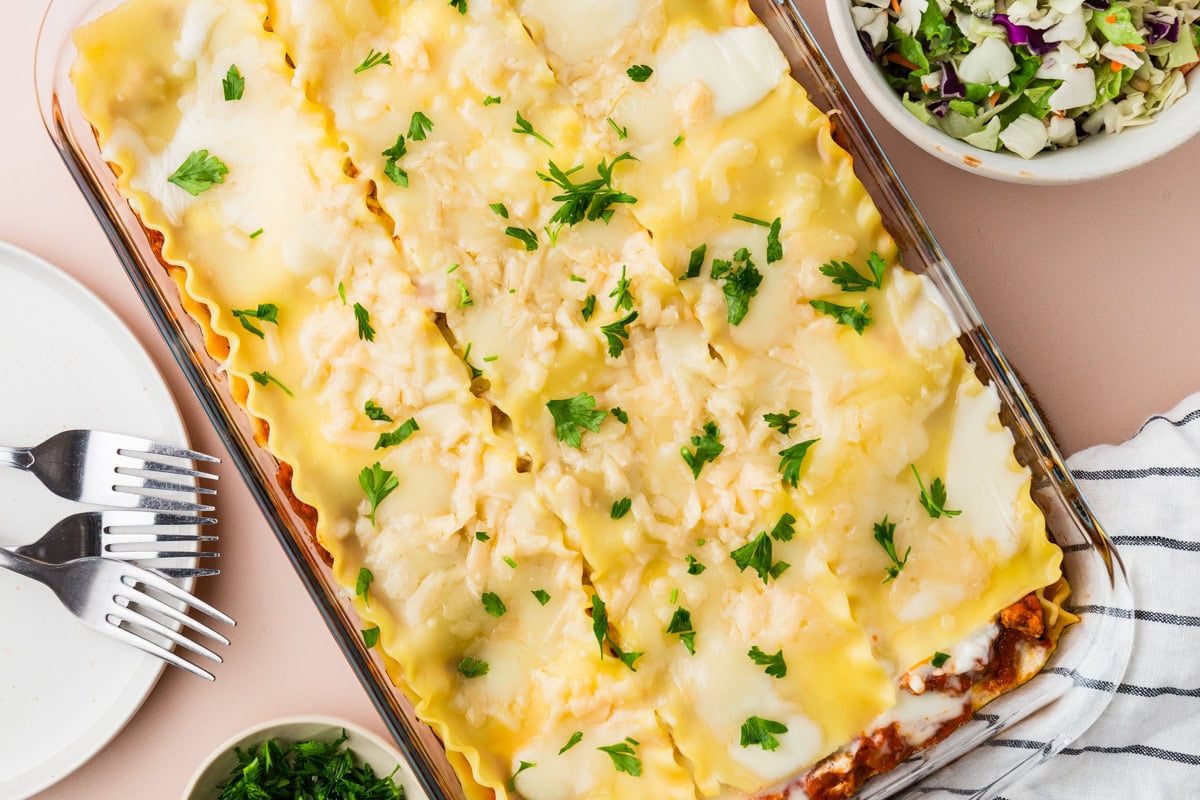 A glass baking dish filled with cheesy Ground Turkey Lasagna, topped with melted cheese and chopped parsley, sits beside a bowl of salad, a striped kitchen towel, plates, and forks on a light pink surface.