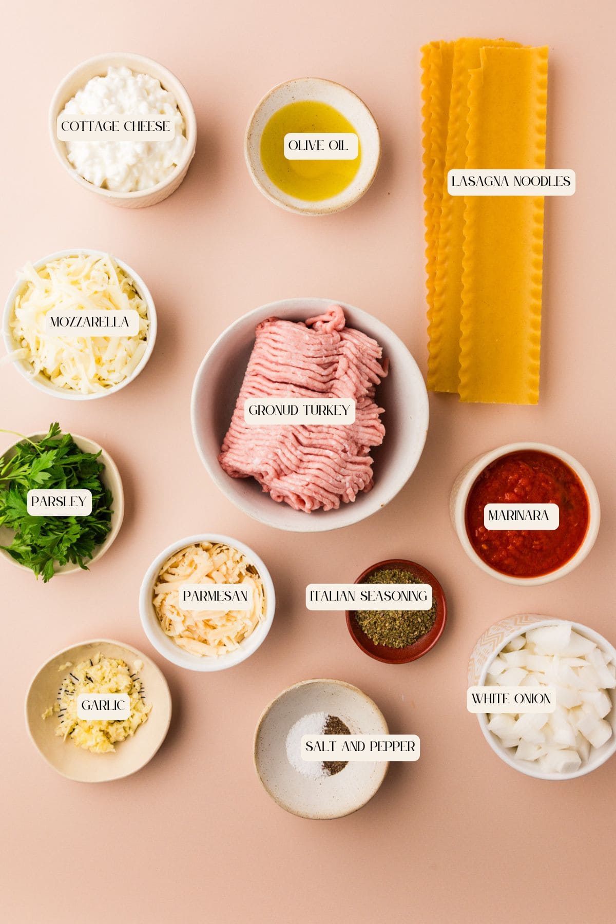 Flat lay of labeled Ground Turkey Lasagna ingredients in bowls: cottage cheese, olive oil, lasagna noodles, mozzarella, ground turkey, marinara sauce, parsley, parmesan, Italian seasoning, garlic, white onion, salt and pepper.