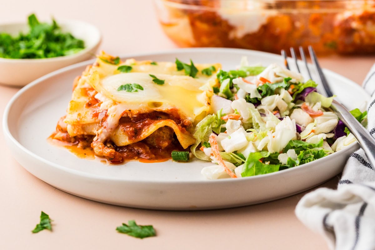 A plate with a serving of Ground Turkey Lasagna and a side of fresh green salad. A fork and napkin are next to the plate, with a dish of chopped herbs and a casserole dish visible in the background.