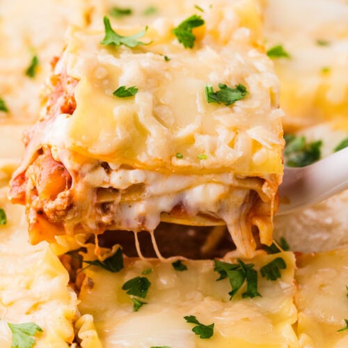 A close-up of a slice of cheesy Ground Turkey Lasagna being lifted from a baking dish, with melted cheese stretching and fresh parsley sprinkled on top.