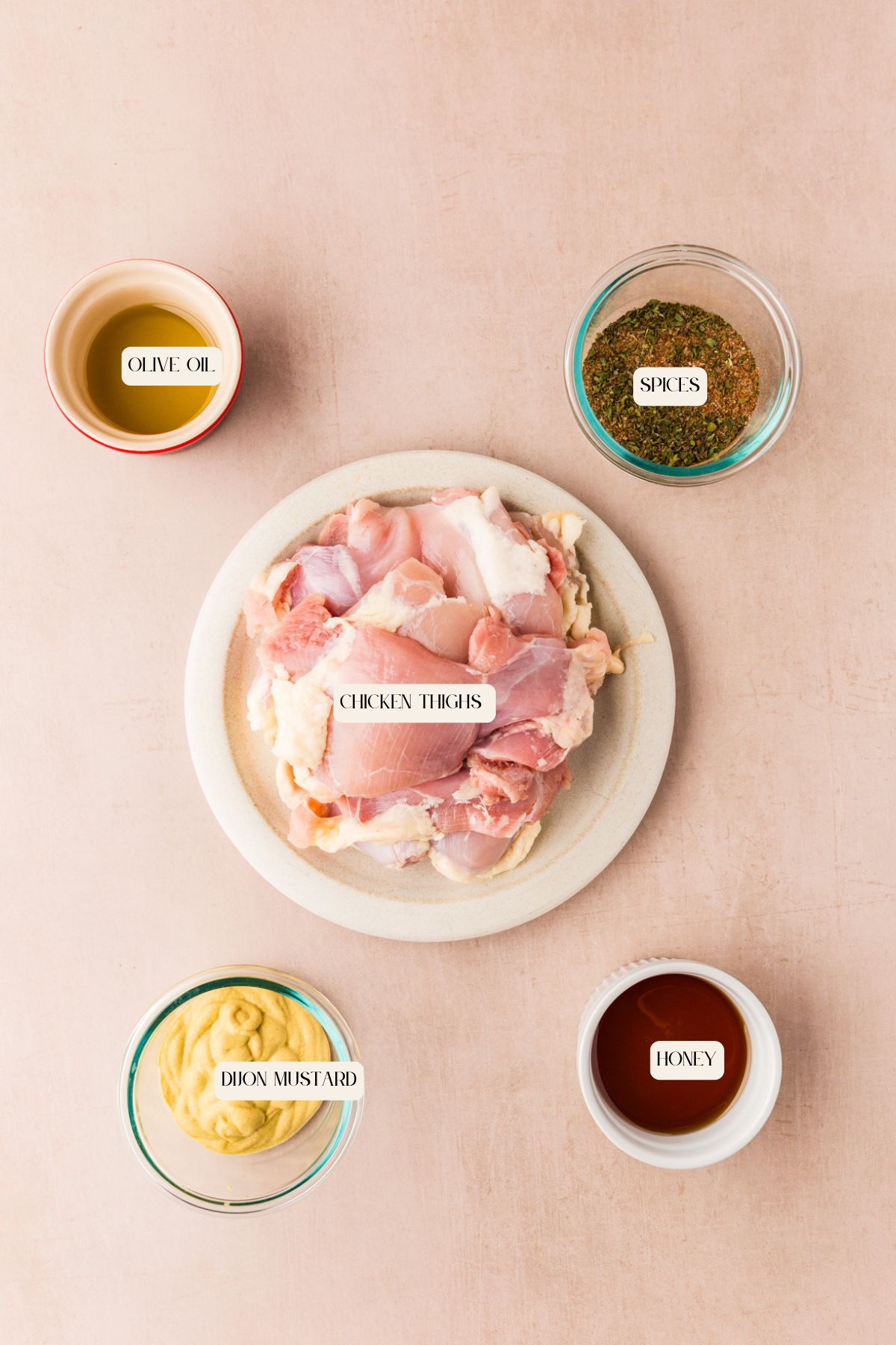 Top-down view of labeled ingredients on a pink surface: raw chicken thighs on a plate in the center, with separate bowls of olive oil, spices, Dijon mustard, and honey arranged around it.