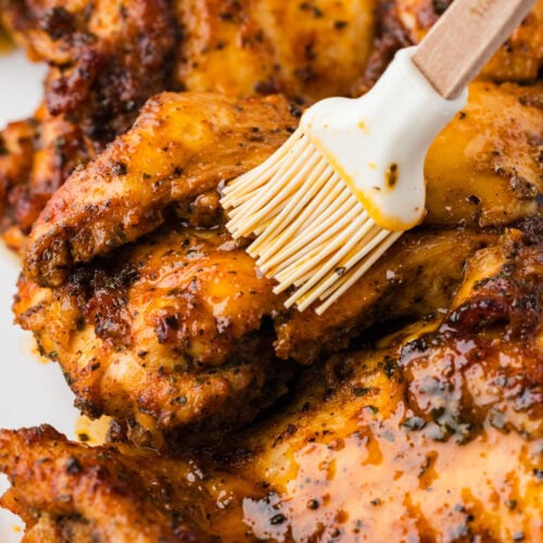 Close-up of grilled chicken pieces being brushed with a glaze or marinade using a silicone brush, showing juicy, golden-brown, and seasoned meat.