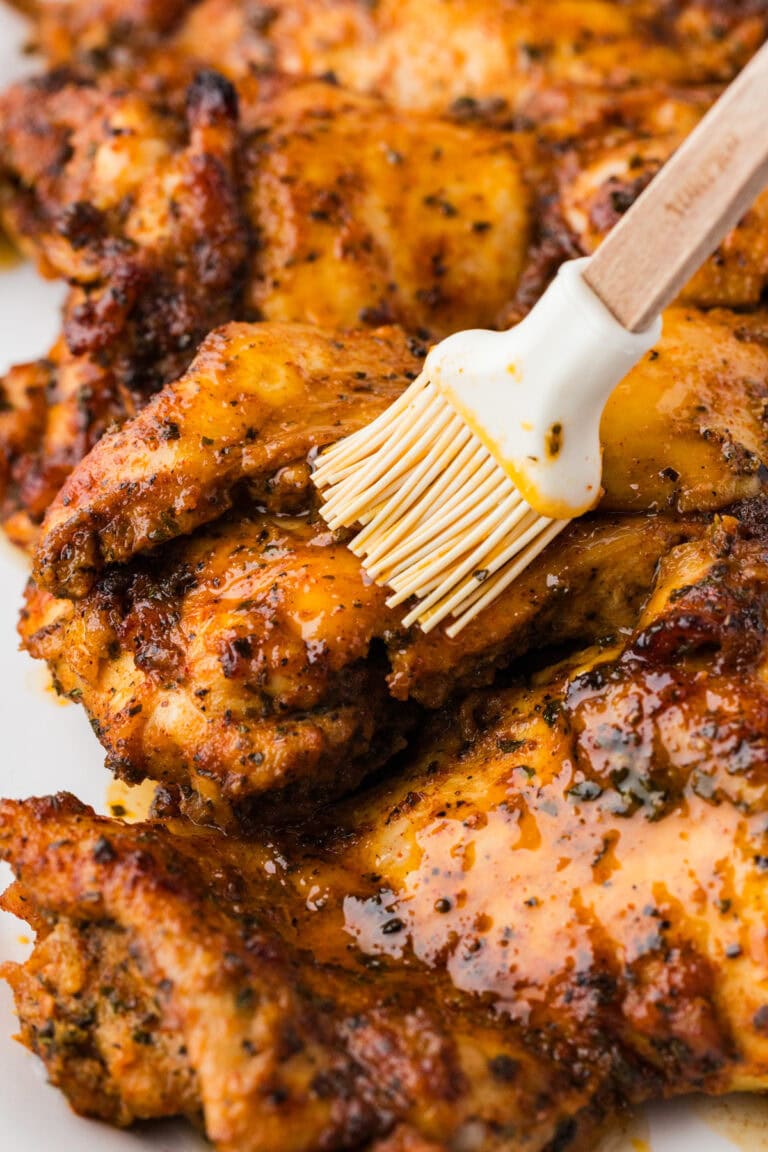 Close-up of grilled chicken pieces being brushed with a glaze or marinade using a silicone brush, showing juicy, golden-brown, and seasoned meat.