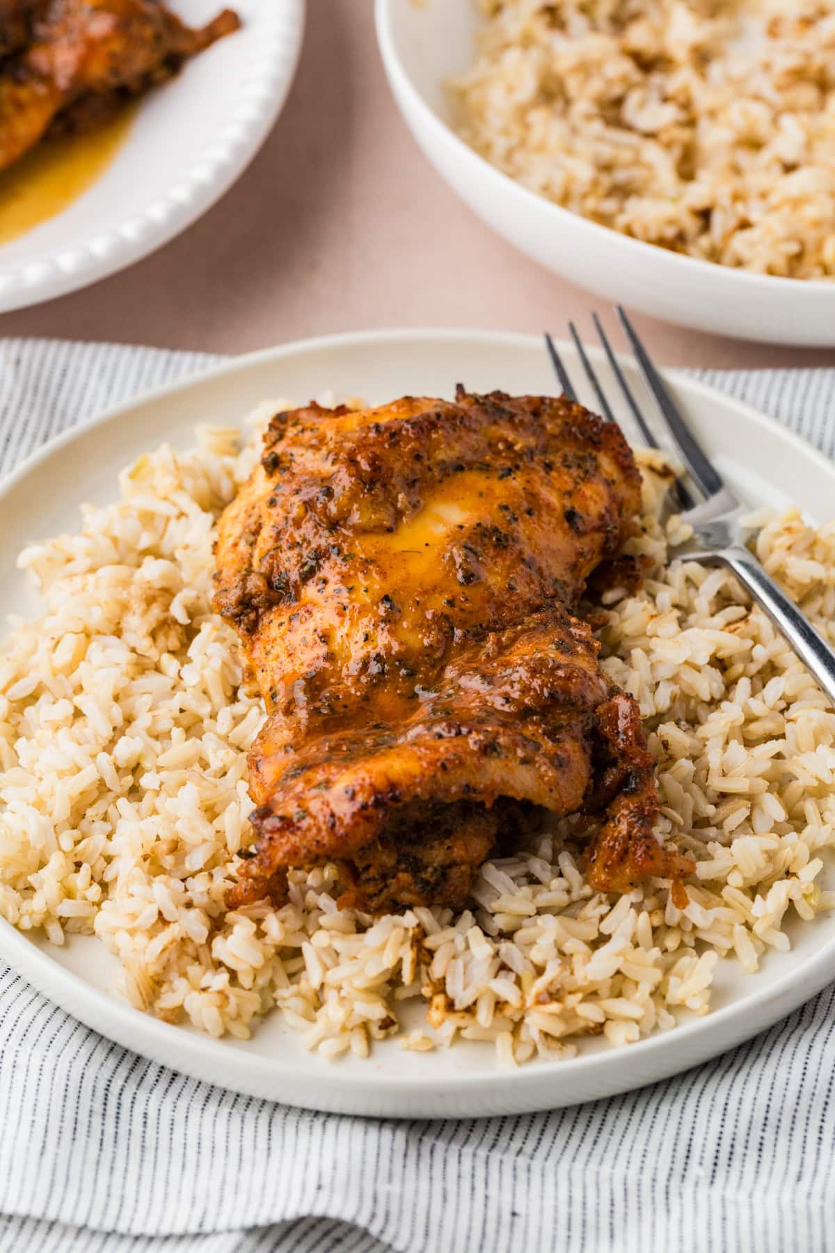A plate of seasoned baked chicken thigh served over a bed of brown rice, with a fork resting on the side. More rice and chicken appear on plates in the background.