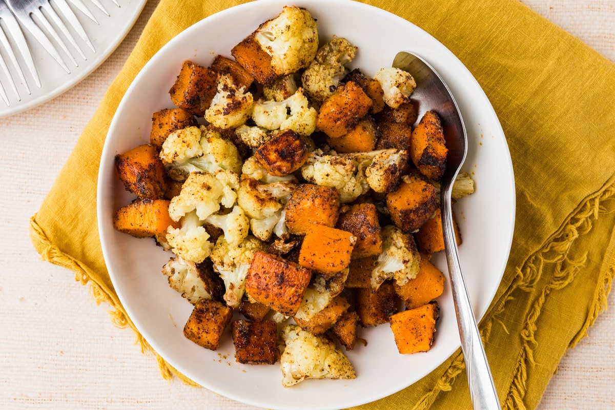 A white bowl filled with roasted cauliflower and sweet potato chunks, seasoned with spices, sits on a yellow cloth napkin. A spoon rests in the bowl, ready for serving.