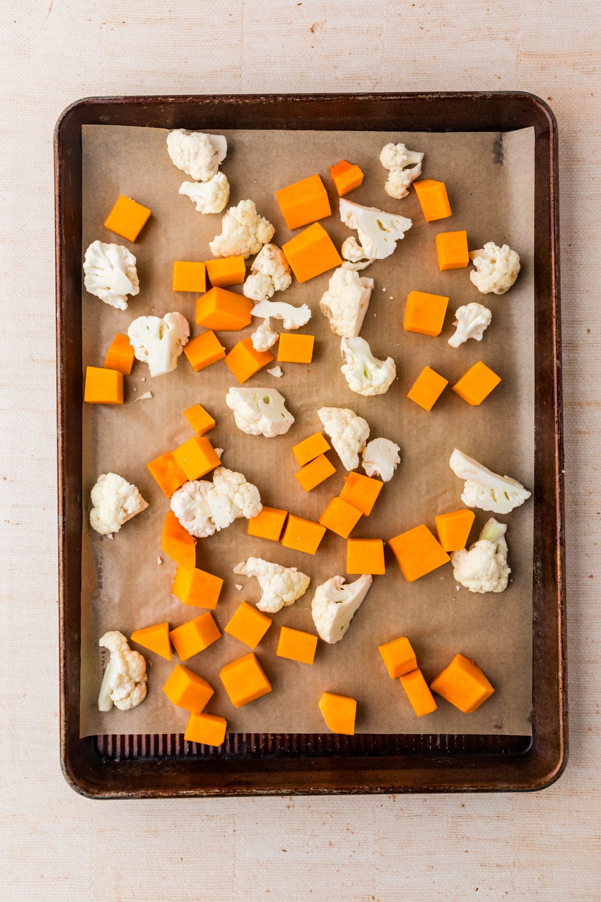 A baking tray lined with parchment paper holds scattered chunks of orange butternut squash and white cauliflower florets, ready for roasting.