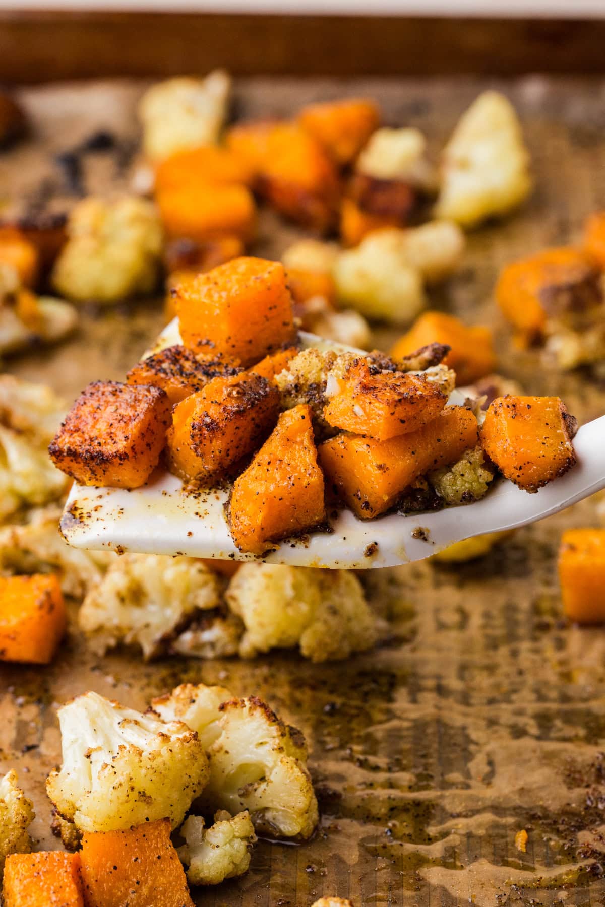 A close-up of a spoon holding roasted cubes of seasoned sweet potatoes and cauliflower, with more roasted vegetables scattered on a baking sheet in the background.