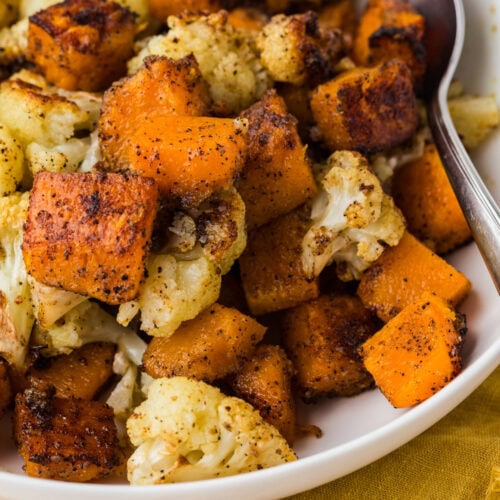 A close-up of a white bowl filled with roasted cauliflower and sweet potato cubes, seasoned with spices. A metal spoon rests on the side of the bowl, which sits on a mustard yellow cloth.