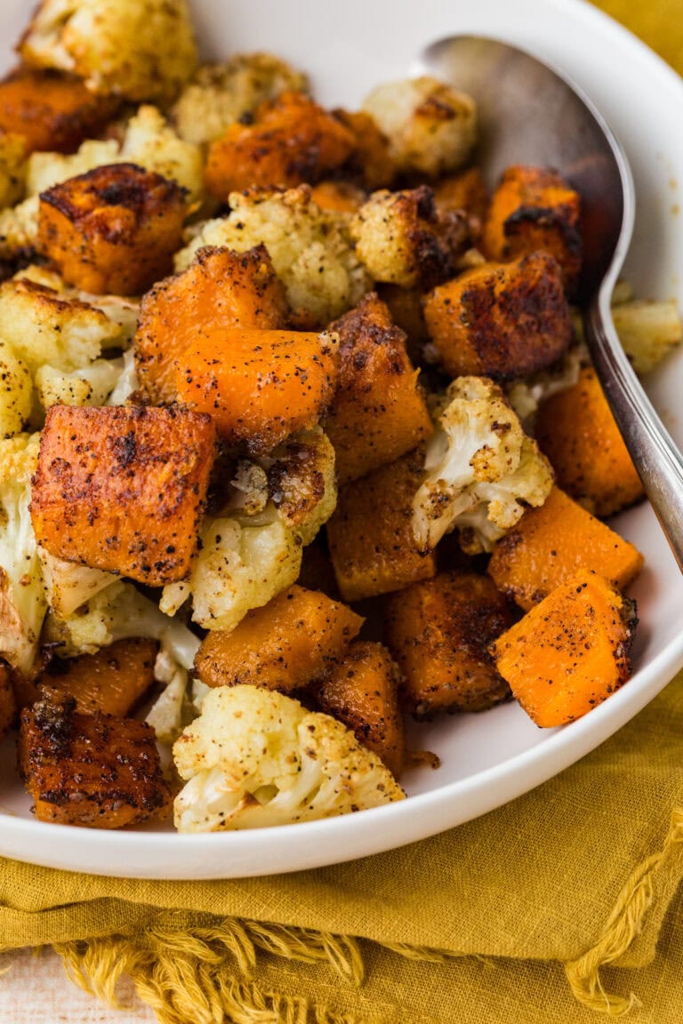 A close-up of a white bowl filled with roasted cauliflower and sweet potato cubes, seasoned with spices. A metal spoon rests on the side of the bowl, which sits on a mustard yellow cloth.