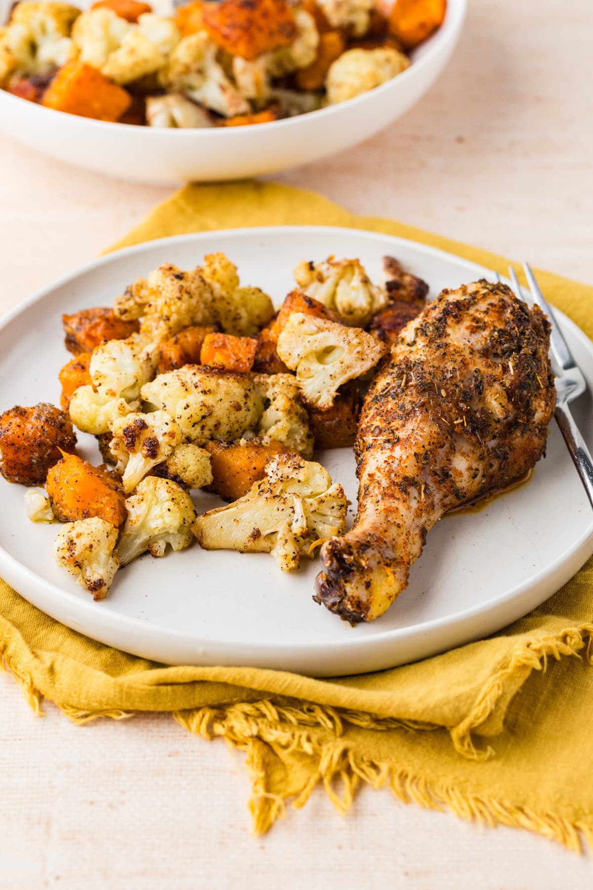 A plate with a seasoned baked chicken drumstick and roasted cauliflower and sweet potatoes, set on a yellow napkin, with a fork on the side and a bowl of more roasted vegetables in the background.