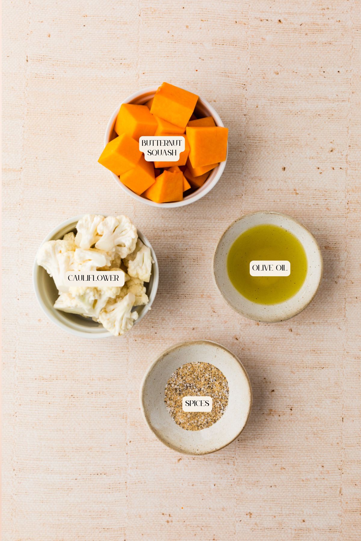 Four small bowls on a beige surface containing labeled ingredients—perfect for making roasted butternut squash and cauliflower—include cubed butternut squash, cauliflower florets, olive oil, and a mix of spices.