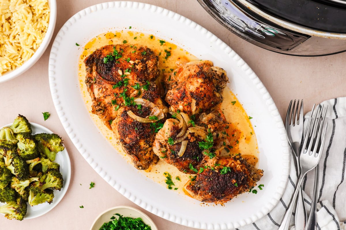 A white platter holds cooked, seasoned chicken thighs topped with herbs and onions in sauce. Its surrounded by a bowl of orzo, a plate of roasted broccoli, chopped herbs, forks, and part of a slow cooker.