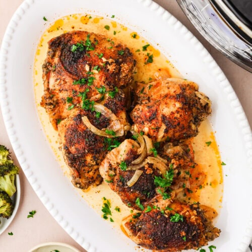 An oval white platter holds five seasoned, browned slow cooker Italian chicken thighs in a creamy sauce, garnished with chopped parsley and sliced onions. A slow cooker, orzo, broccoli, and extra parsley are visible nearby.