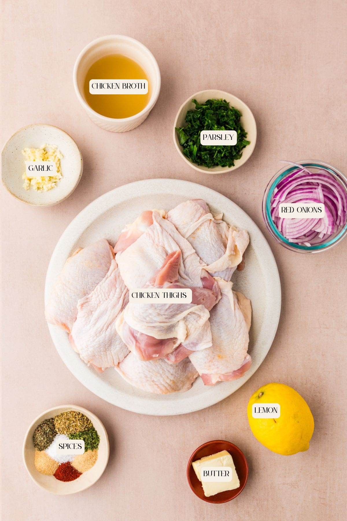 Raw chicken thighs on a plate surrounded by bowls of chopped parsley, sliced red onions, minced garlic, spices, chicken broth, butter, and a whole lemon on a light pink surface. Each ingredient is labeled.