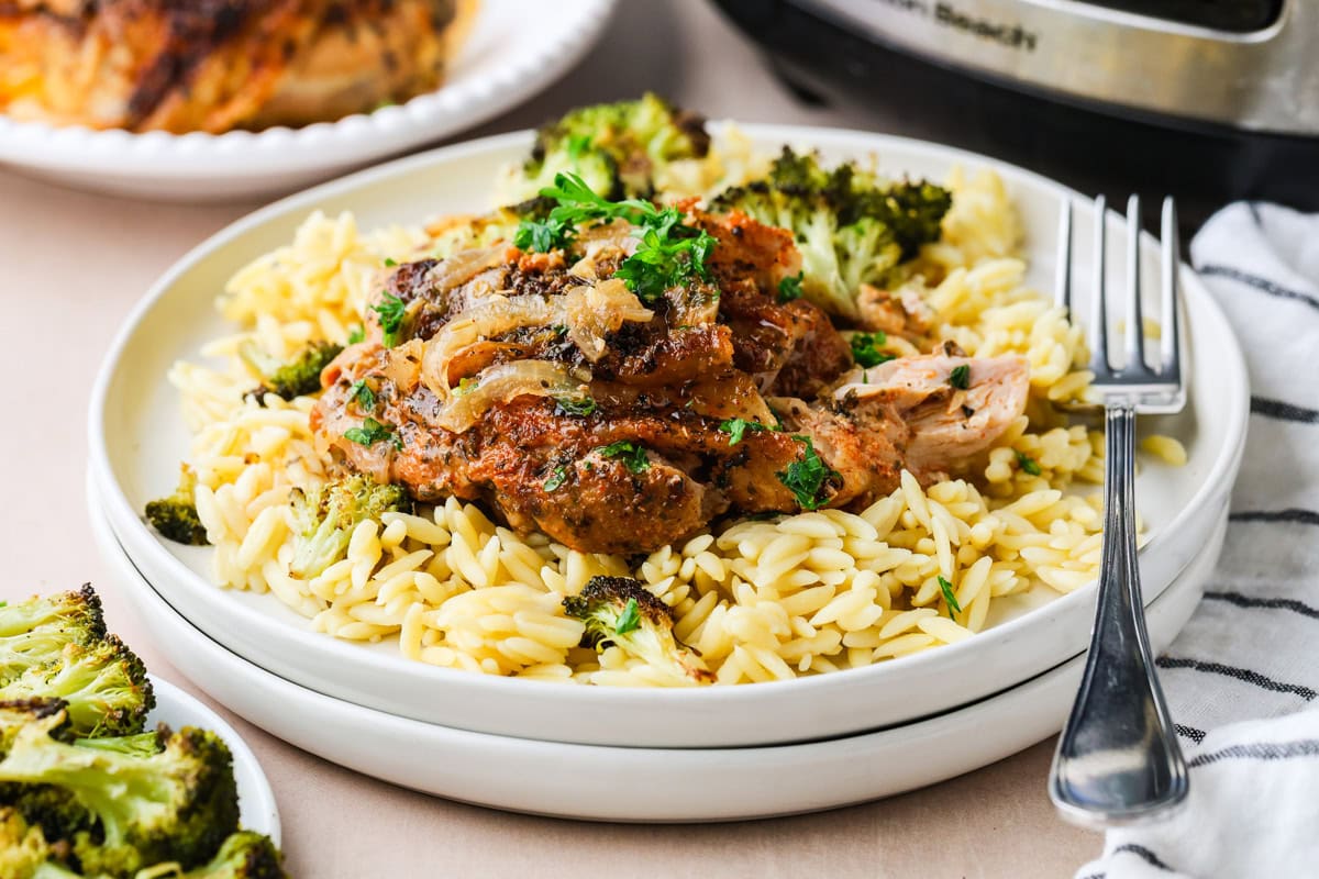 A plate of cooked orzo pasta topped with seasoned, shredded chicken, caramelized onions, fresh parsley, and roasted broccoli, with a fork on the side. In the background are more broccoli and a piece of chicken.