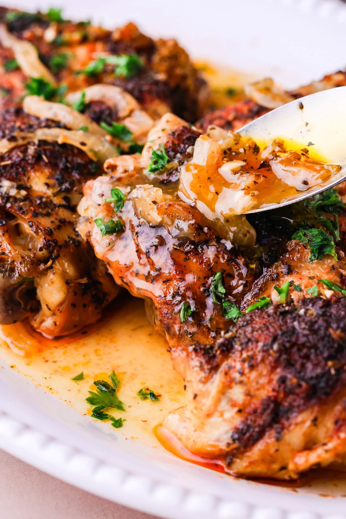 Close-up of seasoned baked chicken breasts topped with sautéed onions and herbs, being drizzled with flavorful pan sauce from a spoon on a white plate.
