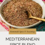 A shallow bowl filled with a Mediterranean Spice Mix and a small brass spoon, set on a colorful patterned cloth. Text overlay reads: “Homemade Spices & Seasonings, MEDITERRANEAN SPICE BLEND, whensaltyandsweetunite.com”.
