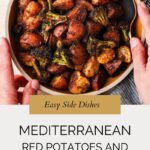 A bowl of roasted red potatoes and broccoli with seasoning sits on a striped cloth. A serving spoon rests in the bowl. Text on the image reads: Mediterranean Roasted Red Potatoes and Broccoli - Easy Side Dishes.
