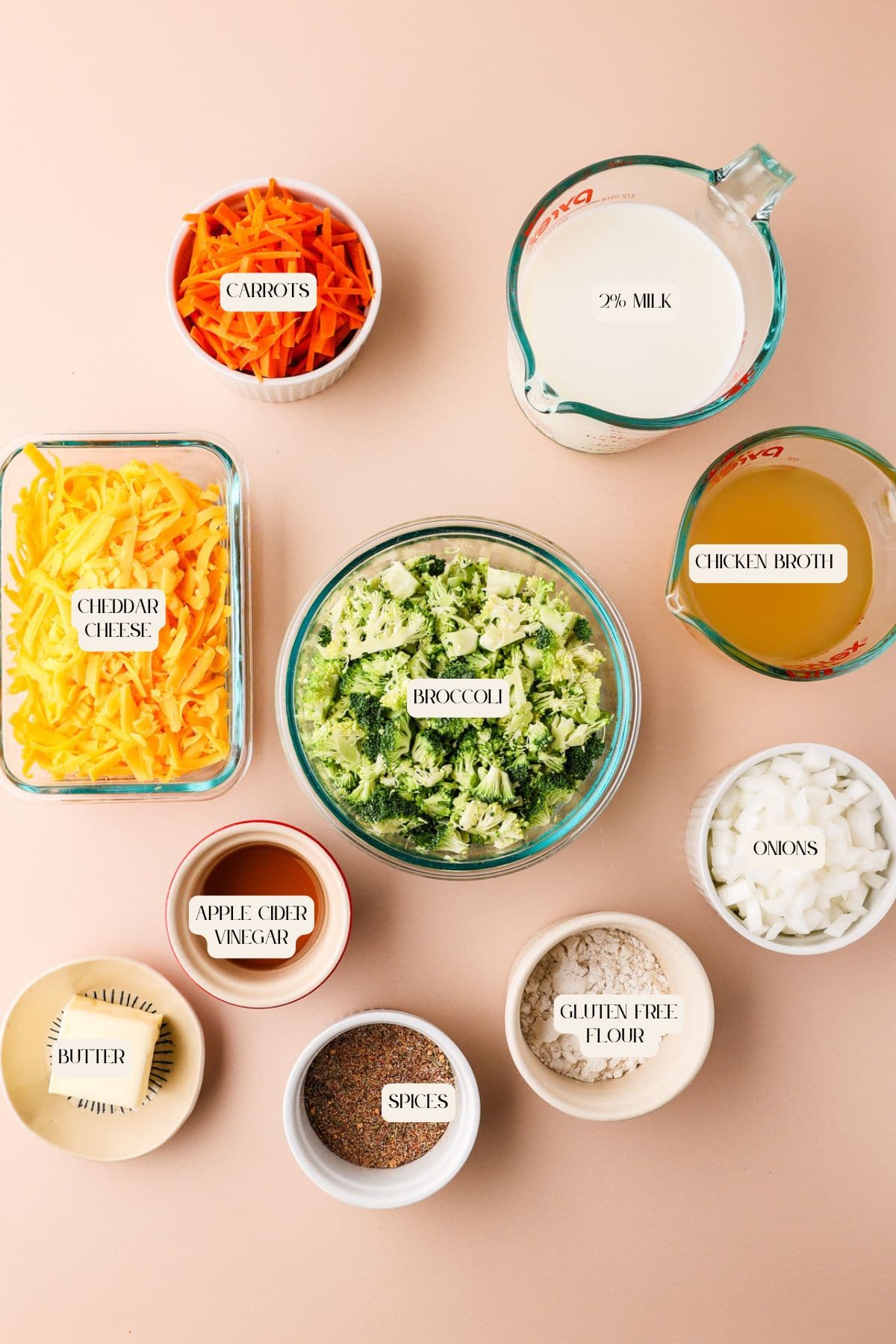 Top-down view of various soup ingredients in bowls: shredded carrots, cheddar cheese, chopped broccoli, milk, chicken broth, diced onions, butter, apple cider vinegar, gluten-free flour, and mixed spices on a beige surface.