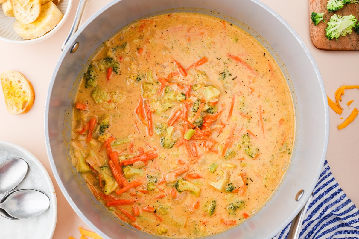 A large pot filled with creamy broccoli and carrot soup. Sliced carrots and broccoli florets are visible in the orange-tinted broth. Slices of bread, spoons, and a napkin are nearby on the table.