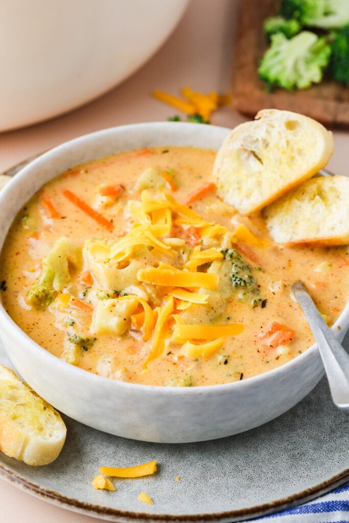A bowl of creamy gluten free broccoli cheddar soup topped with shredded cheddar cheese, served with slices of toasted bread on the side. A spoon rests inside the bowl, and broccoli florets are visible in the background.