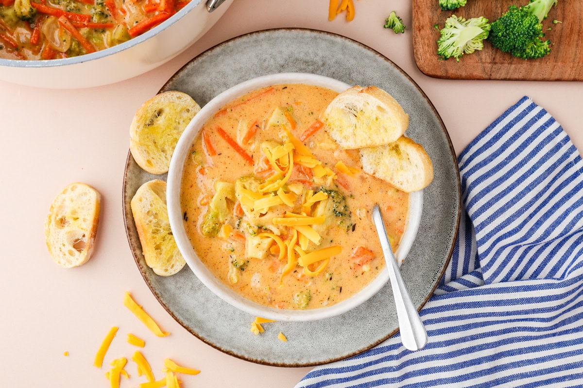 A bowl of creamy broccoli cheddar soup topped with shredded cheddar cheese, served with toasted bread slices on a plate. A striped blue napkin and a cutting board with broccoli are nearby.