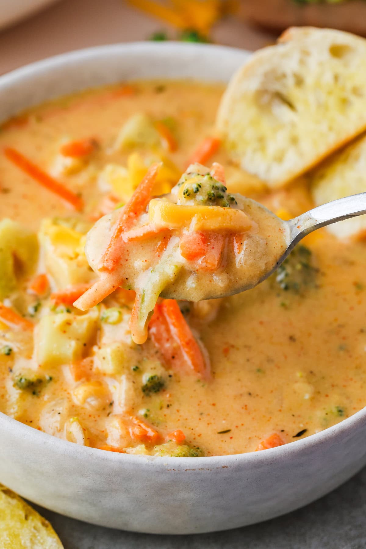 A close-up of creamy gluten free broccoli cheddar soup with carrots and cheese in a white bowl, a spoon lifting a bite, and slices of bread on the side.