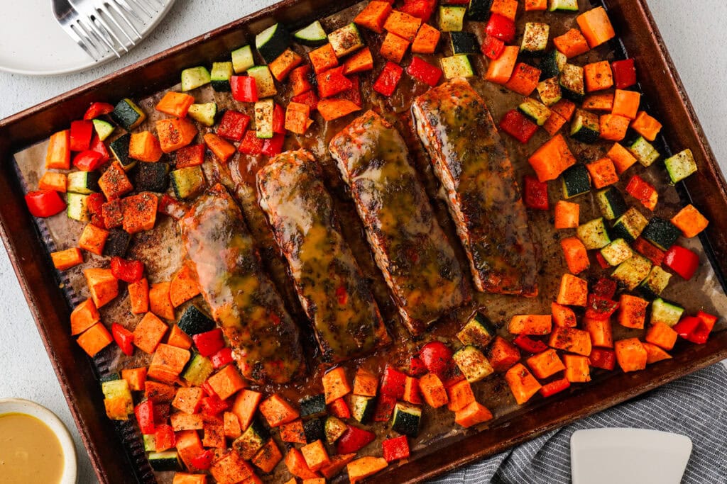 A sheet pan with four glazed salmon fillets surrounded by roasted vegetables, including diced sweet potatoes, zucchini, and red bell peppers. Plates, a fork, and a small bowl of sauce are visible nearby.