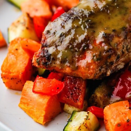 A close-up of a glazed, cooked chicken breast served on a white plate with roasted vegetables, including sweet potatoes, red bell peppers, and zucchini.