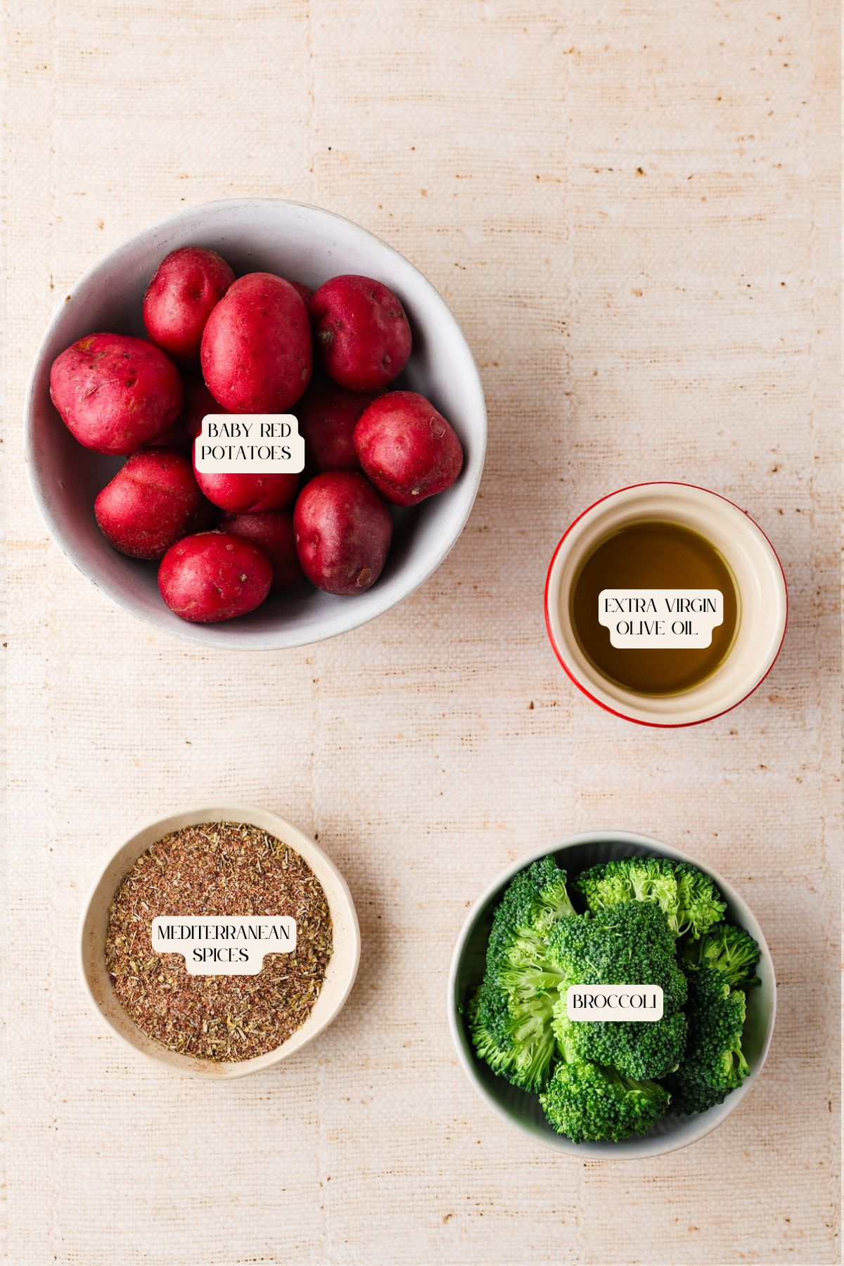Four bowls contain ingredients for roasted red potatoes and broccoli: baby red potatoes, extra virgin olive oil, Mediterranean spices, and broccoli. Each bowl is labeled and arranged on a beige surface.