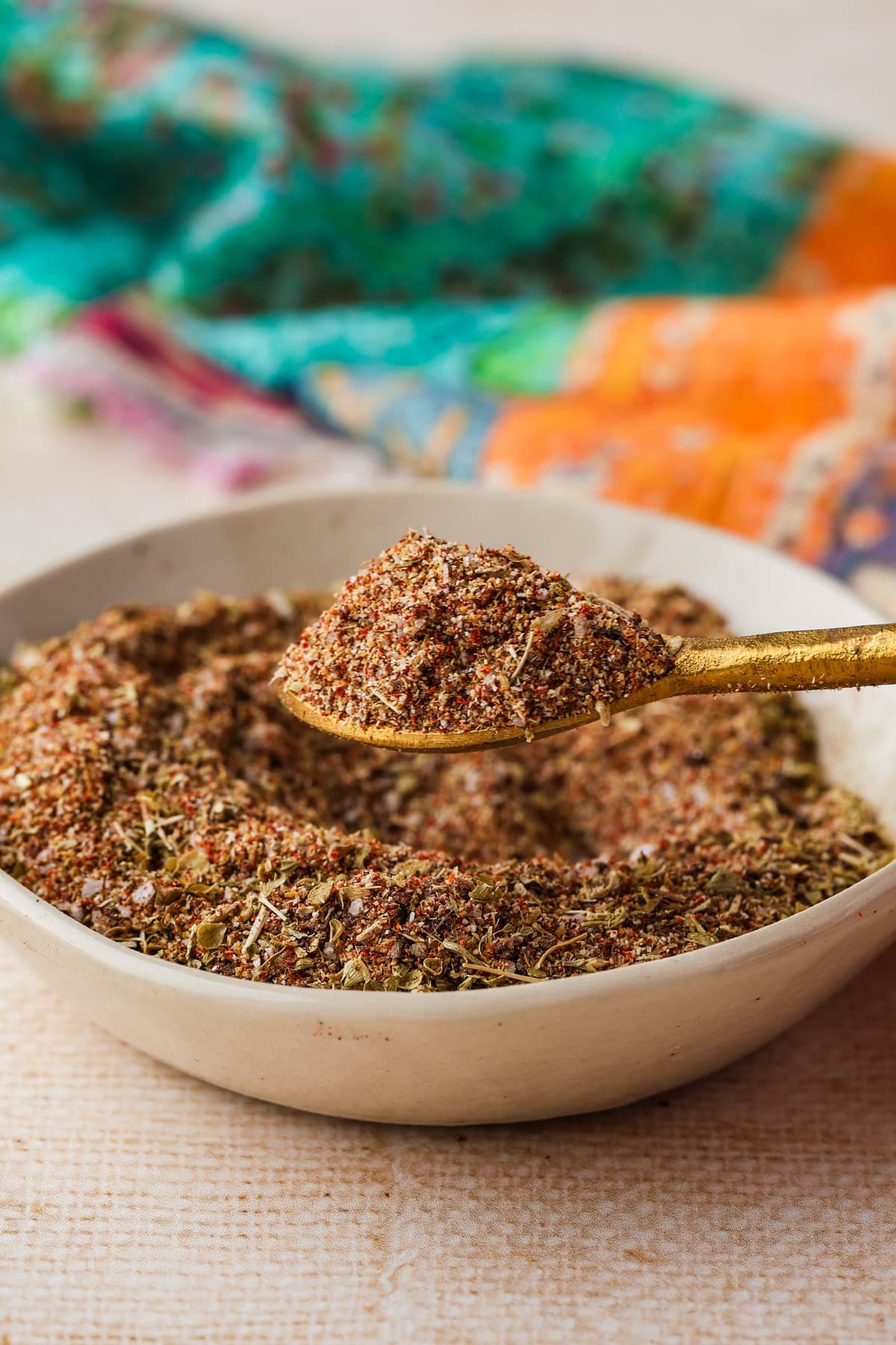 A gold spoon holding a blend of mixed spices over a white bowl filled with the same spice mix, with a colorful cloth in the background.