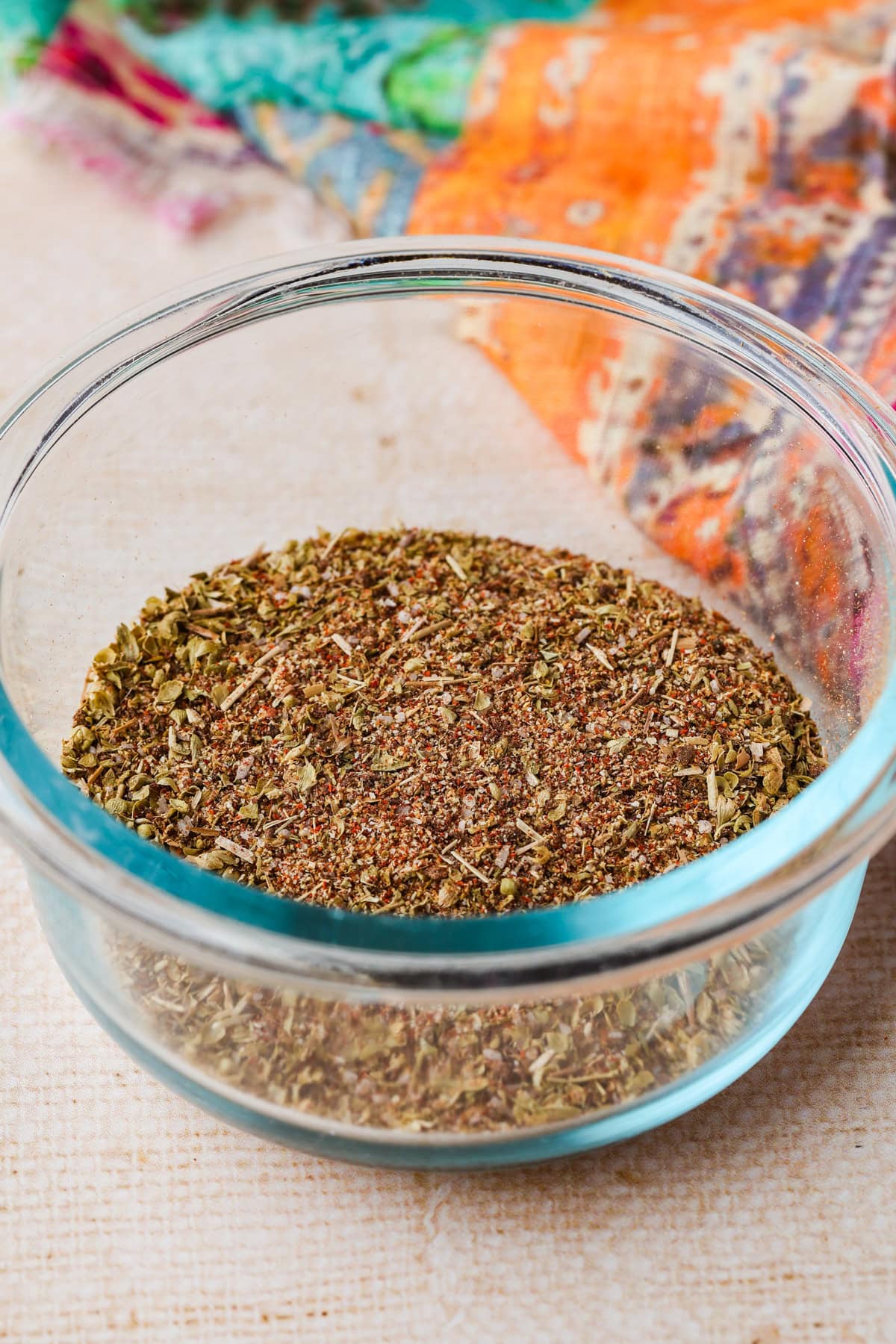 A clear glass bowl filled with a mixture of dried spices and herbs sits on a light textured surface, with a colorful fabric partially visible in the background.