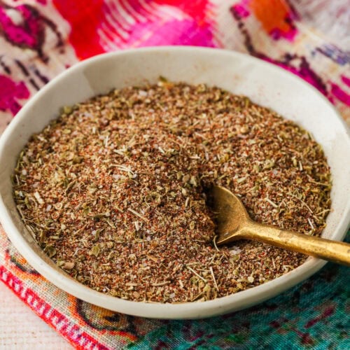 A small white bowl filled with a coarse, mixed spice blend sits on a colorful, patterned cloth, with a gold spoon resting in the bowl.