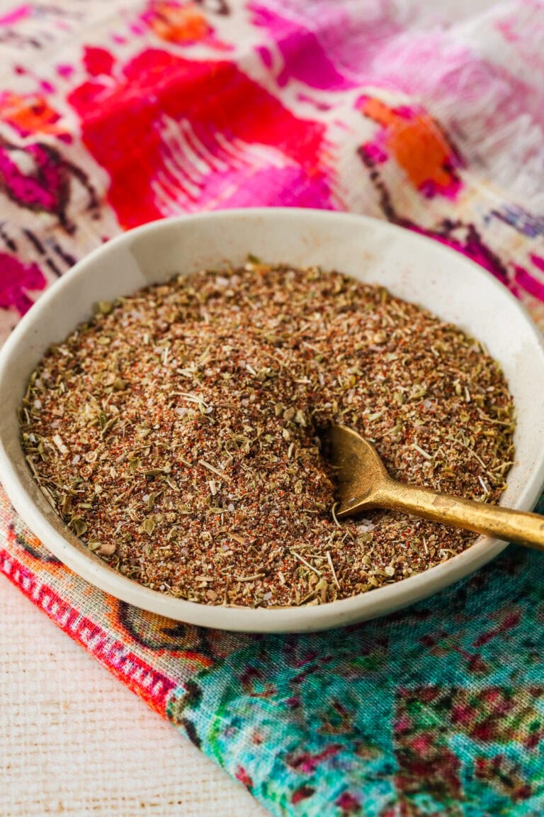 A small white bowl filled with a coarse, mixed spice blend sits on a colorful, patterned cloth, with a gold spoon resting in the bowl.
