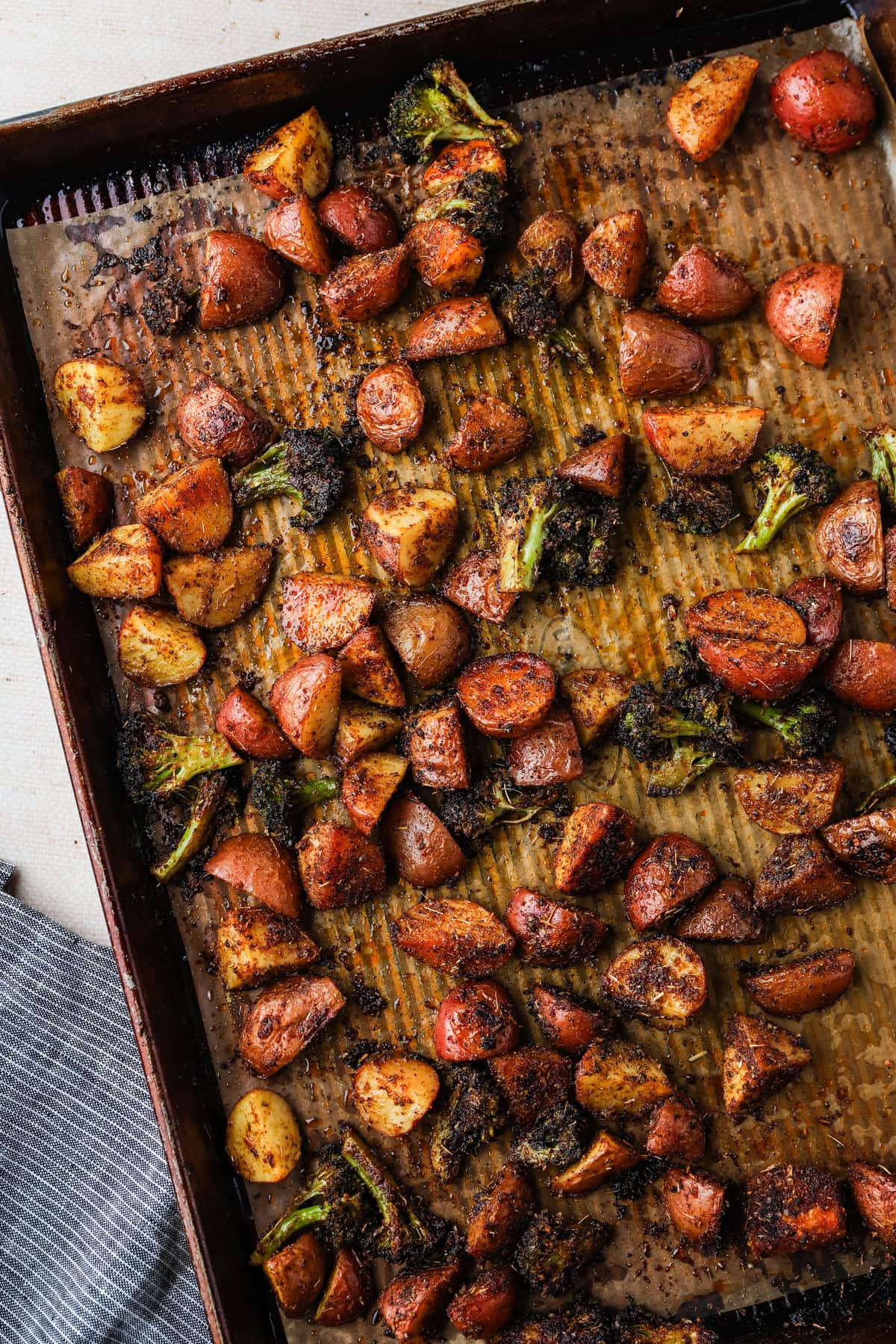 A baking tray lined with parchment paper holds roasted red potatoes and broccoli, seasoned to perfection and browned, spread evenly across the surface. A gray striped napkin is partially visible in the bottom left corner.
