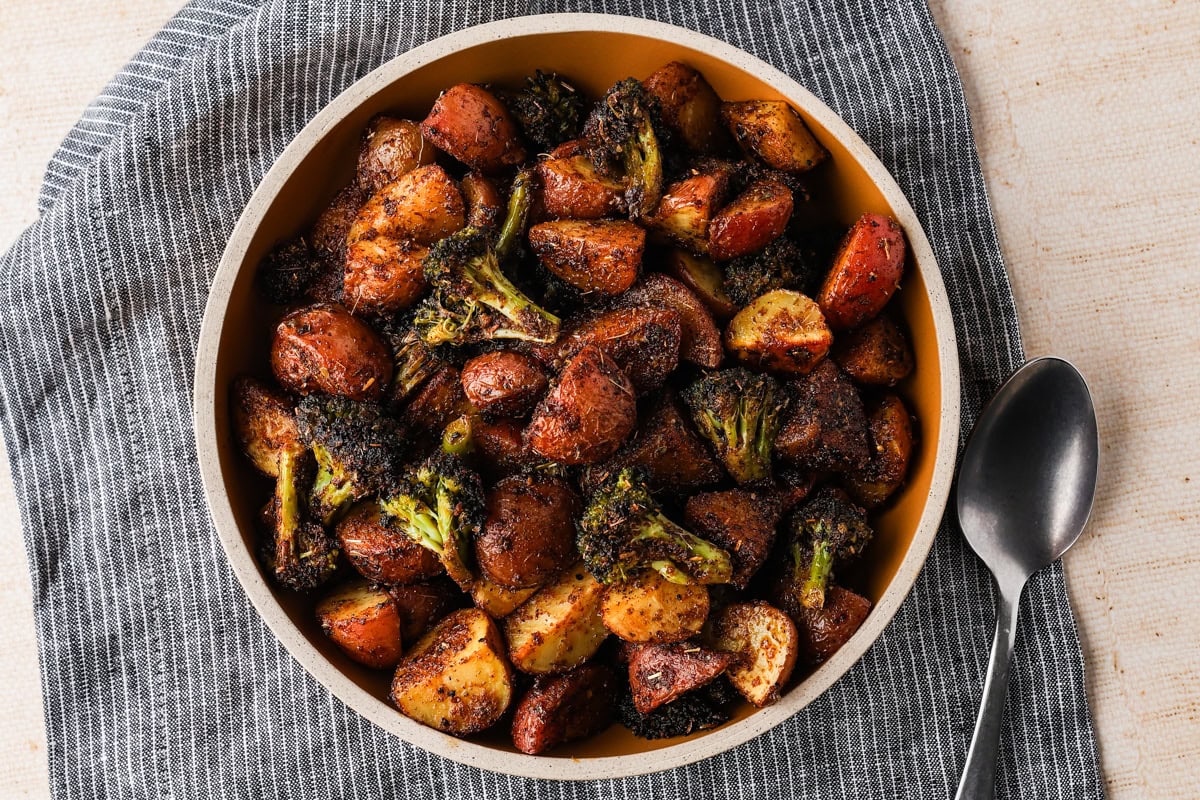 A bowl of Mediterranea roasted potatoes and broccoli, seasoned with spices, sits on a gray-striped cloth beside a spoon.