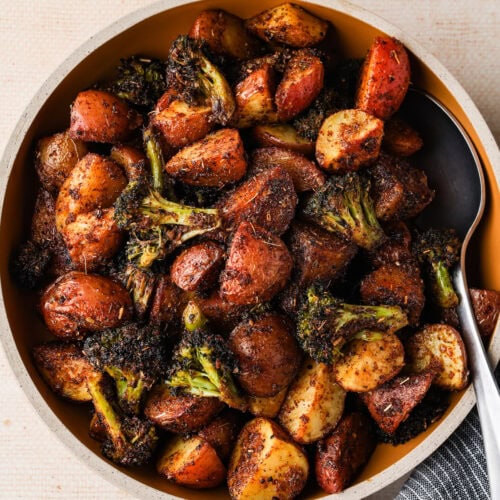 A bowl filled with roasted red potatoes and broccoli, seasoned with spices, sits on a light-colored surface beside a dark-striped napkin. A serving spoon rests inside the bowl.