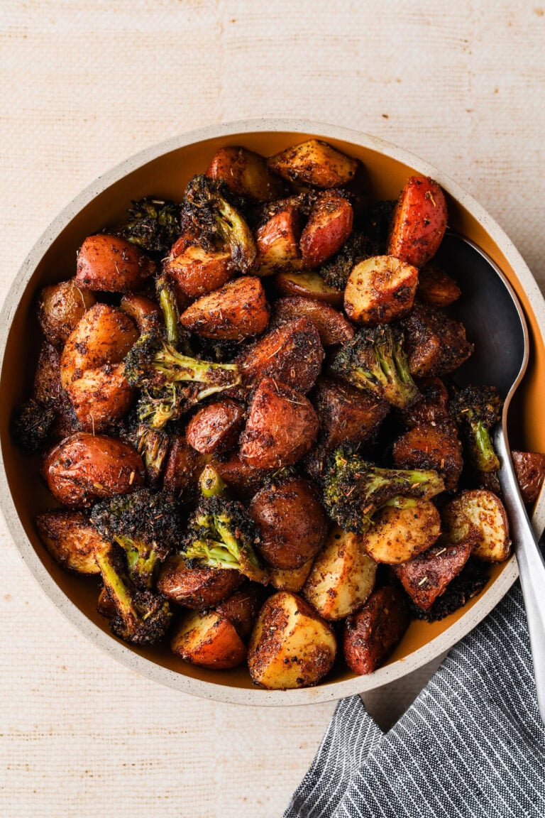 A bowl filled with roasted red potatoes and broccoli, seasoned with spices, sits on a light-colored surface beside a dark-striped napkin. A serving spoon rests inside the bowl.