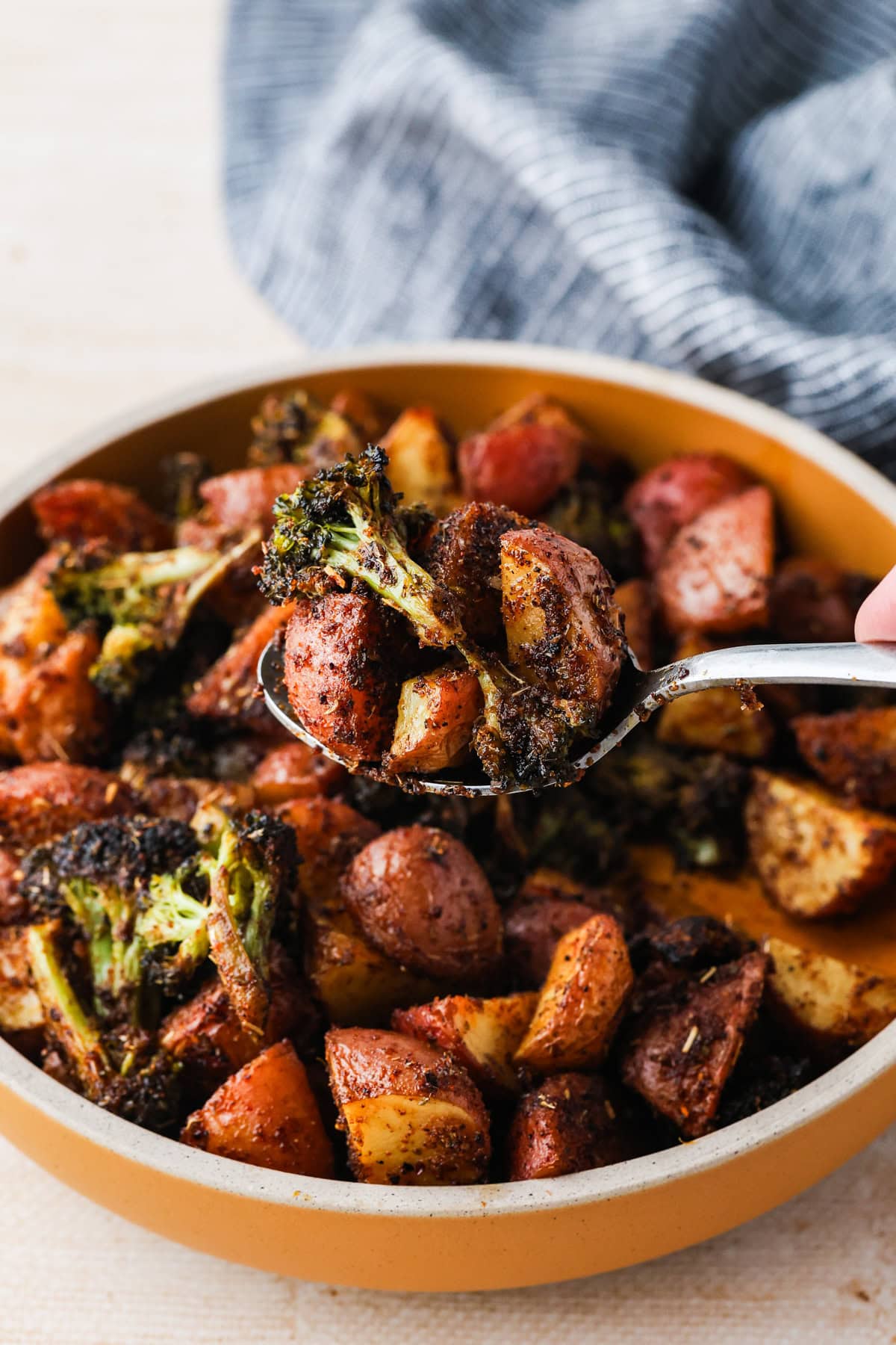 A close-up of a spoonful of roasted red potatoes and broccoli, lifted from a pan filled with more, all seasoned with herbs. A blue-and-white cloth is visible in the background.