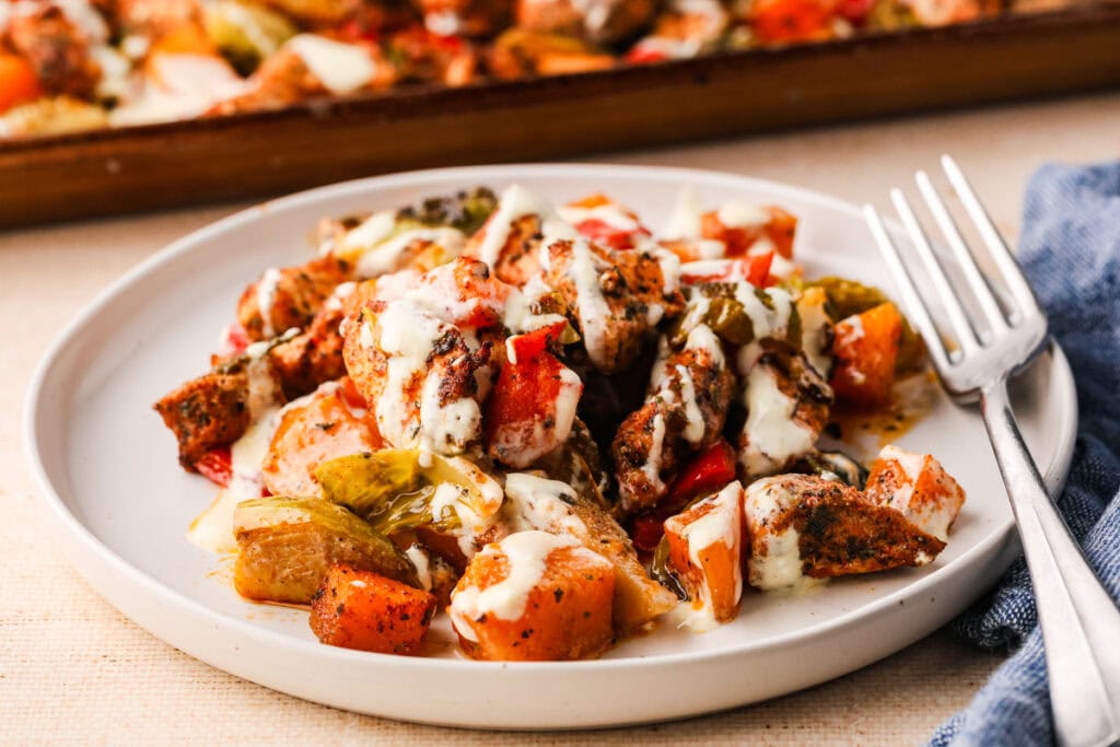 A white plate holds roasted vegetables and sheet pan chicken breast pieces drizzled with a creamy white sauce. A fork sits beside the plate, and a blue napkin is nearby.